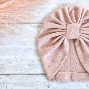 May include: A pale pink baby turban hat with a bow feature. The hat is made from a soft, breathable material with a delicate openwork pattern. The hat is designed to fit a baby's head and is a fashionable accessory.