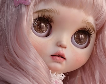 Può includere: Primo piano di una bambola con capelli rosa e grandi occhi marroni. La bambola ha lunghe ciglia, guance rosee e un delicato colore delle labbra rosa. Due fermagli per capelli a farfalla, uno rosa e uno blu, adornano i capelli della bambola. La bambola indossa un vestito rosa con un colletto di pizzo.
