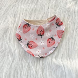 May include: A baby bib with a pink background featuring a pattern of smiling strawberries, white flowers, and small pink stars. The bib has a rounded shape and a soft, neutral-colored lining.