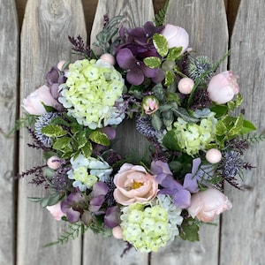 May include: A circular floral wreath featuring a variety of flowers in shades of pink, purple, and green. The wreath includes hydrangeas, roses, and other greenery, creating a full and textured appearance. The background is a weathered wooden surface.