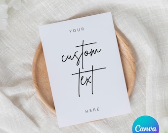 May include: A white card with the words "YOUR custom text HERE" in black script. The card is on a wooden plate, set against a white fabric background. The Canva logo is in the bottom right corner.