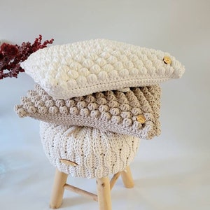May include: A stack of three crocheted cushions in white and beige with wooden buttons on a wooden stool. The cushions are made with a textured bobble stitch.