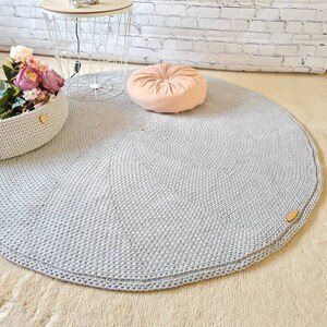 May include: A round, gray, crocheted rug with a light pink velvet cushion on top. The rug is on a light beige surface.