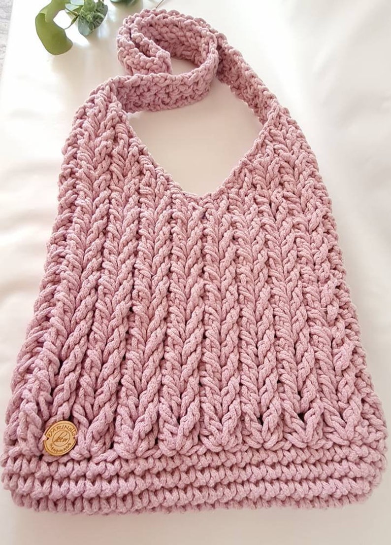 Puede incluir: Un bolso de hombro de crochet rosa con una correa trenzada y una etiqueta de madera que dice "Handmade with Love".