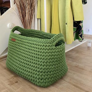 Peut inclure: Un grand panier rectangulaire crocheté à la main, de couleur verte vive. Le panier est doté de poignées robustes et d'un tissage épais et texturé. Une petite étiquette en cuir marron est attachée sur le côté. Le panier est conçu pour le rangement.