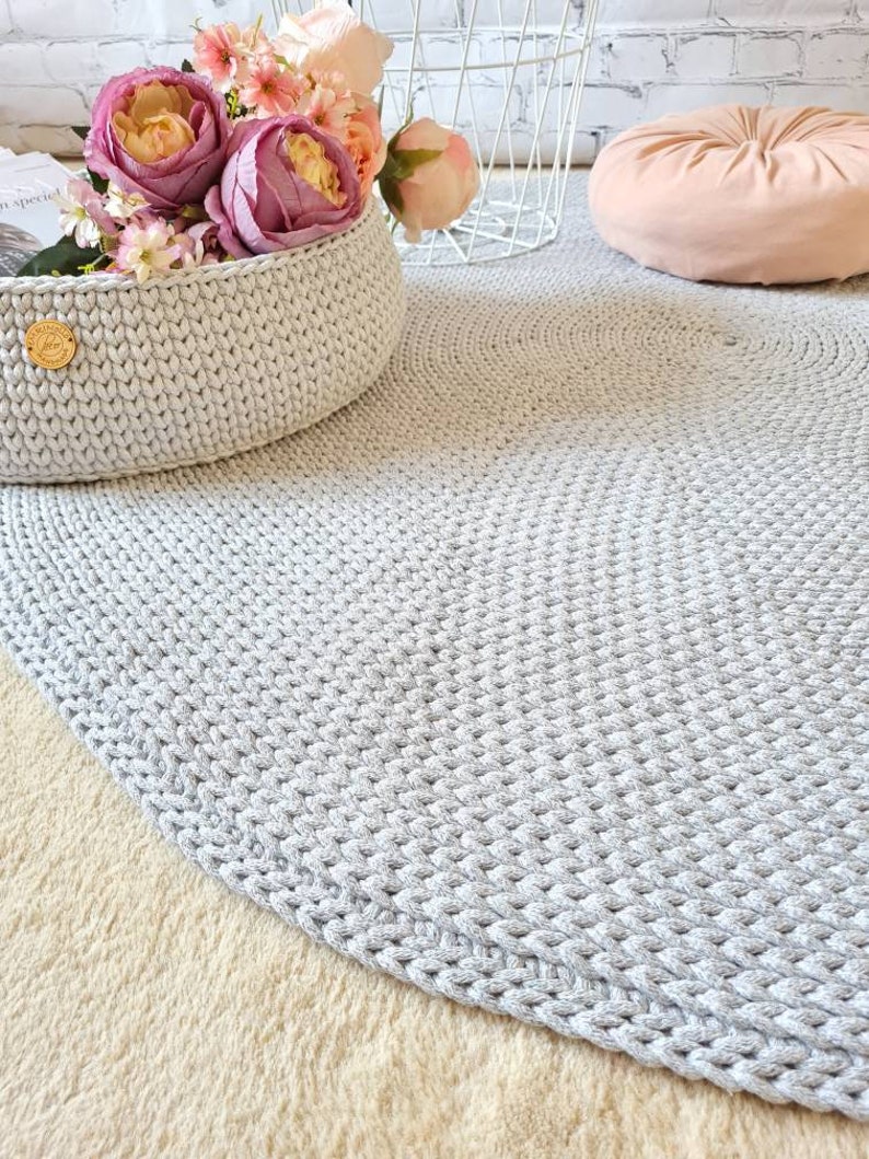 May include: A gray crocheted round rug with a basket of pink flowers in the background.