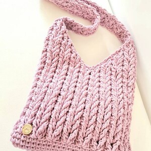 Puede incluir: Un bolso de hombro de crochet de color morado claro con una correa trenzada y un peque&ntilde;o bot&oacute;n dorado.