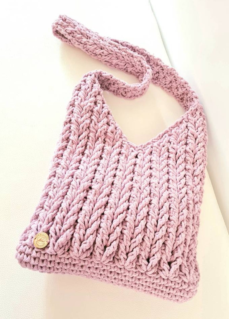 Puede incluir: Un bolso de hombro de crochet de color morado claro con una correa trenzada y un peque&ntilde;o bot&oacute;n dorado.