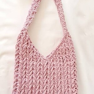Puede incluir: Un bolso de hombro de crochet rosa con una correa trenzada y un peque&ntilde;o bot&oacute;n dorado.