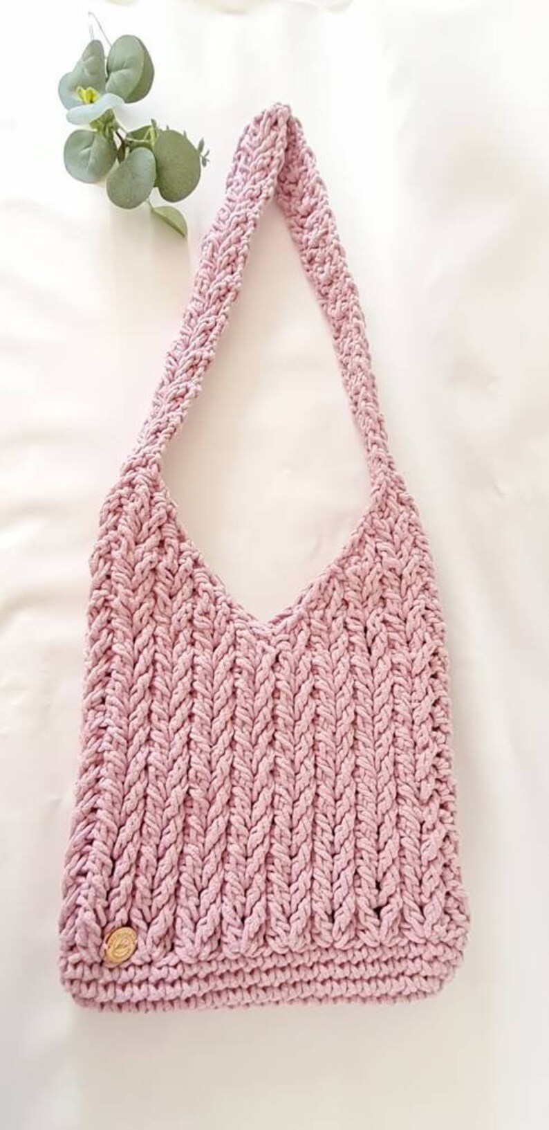Puede incluir: Un bolso de hombro de crochet rosa con una correa trenzada y un peque&ntilde;o bot&oacute;n dorado.
