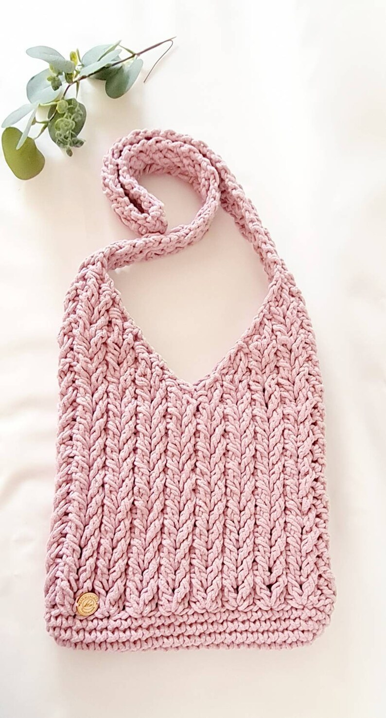 Puede incluir: Un bolso de hombro de crochet rosa con una correa trenzada. El bolso tiene un peque&ntilde;o bot&oacute;n dorado en la parte inferior.
