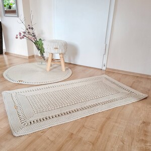 May include: Two crocheted rugs in a cream color. The larger rug is rectangular and the smaller rug is round. A wooden stool with a crocheted cushion is in the background.