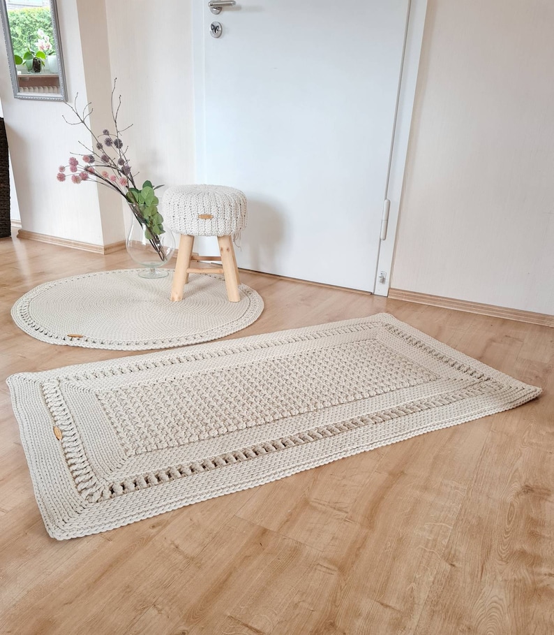 May include: Two crocheted rugs in a cream color. The larger rug is rectangular and the smaller rug is round. A wooden stool with a crocheted cushion is in the background.