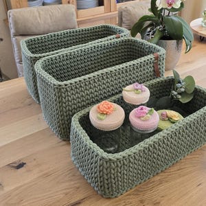 May include: Three green crocheted storage baskets of varying sizes, stacked on top of each other. The smallest basket contains four small glass jars with crocheted lids, decorated with colourful flowers.