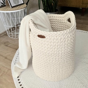 Op de afbeelding: Crèmekleurige, handgebreide opbergmand met oorvormige handvatten. Een zachte, crèmekleurige deken hangt over de rand. De mand heeft een klein, bruin leren label. De mand staat op een wit, rond tapijt.