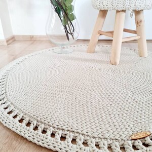 May include: A round, beige crocheted rug with a decorative border. The rug is on a wooden floor and a small wooden stool with a crocheted seat is in the background.