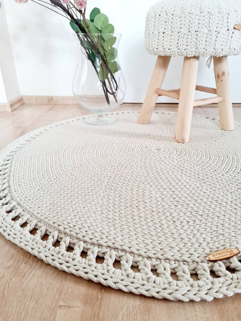May include: A round, beige crocheted rug with a decorative border. The rug is on a wooden floor and a small wooden stool with a crocheted seat is in the background.