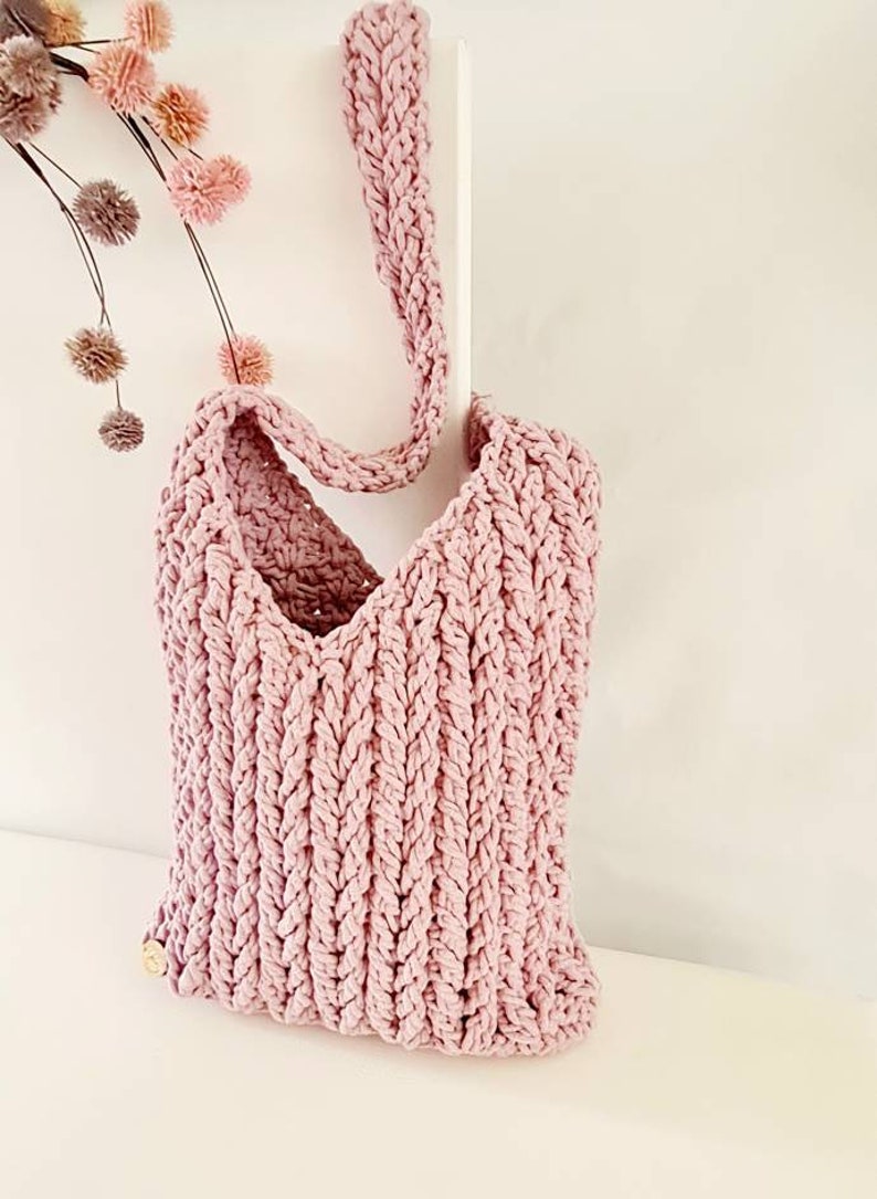 Puede incluir: Una bolsa de mano de crochet rosa con una correa larga. La bolsa tiene un dise&ntilde;o simple y texturizado.