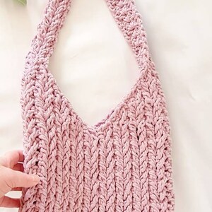 Puede incluir: Un bolso de mano de crochet rosa con una correa trenzada y un peque&ntilde;o bot&oacute;n redondo dorado.