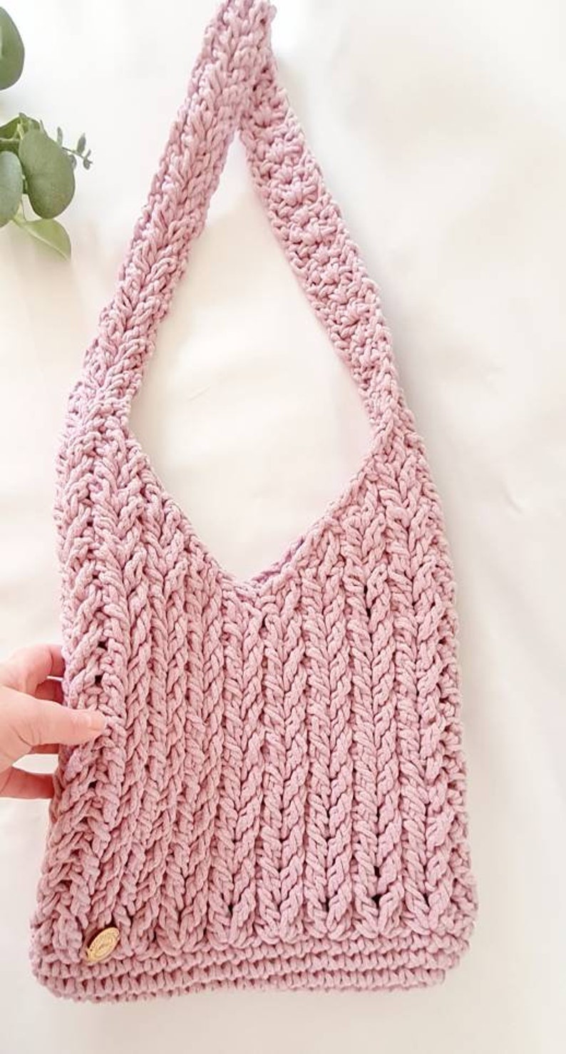 Puede incluir: Un bolso de mano de crochet rosa con una correa trenzada y un peque&ntilde;o bot&oacute;n redondo dorado.