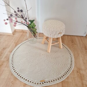 May include: A round, cream-colored crocheted rug with a decorative border. A small wooden stool with a cream-colored crocheted cushion sits on the rug.