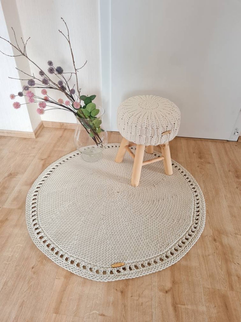May include: A round, cream-colored crocheted rug with a decorative border. A small wooden stool with a cream-colored crocheted cushion sits on the rug.