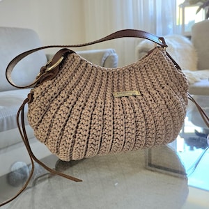 Könnte beinhalten: Eine hellbraune Häkeltasche mit braunen Lederriemen und -details. Die Tasche hat eine geschwungene Form und ein kleines goldfarbenes Metallschild. Der Riemen ist mit einer goldfarbenen Schnalle befestigt.