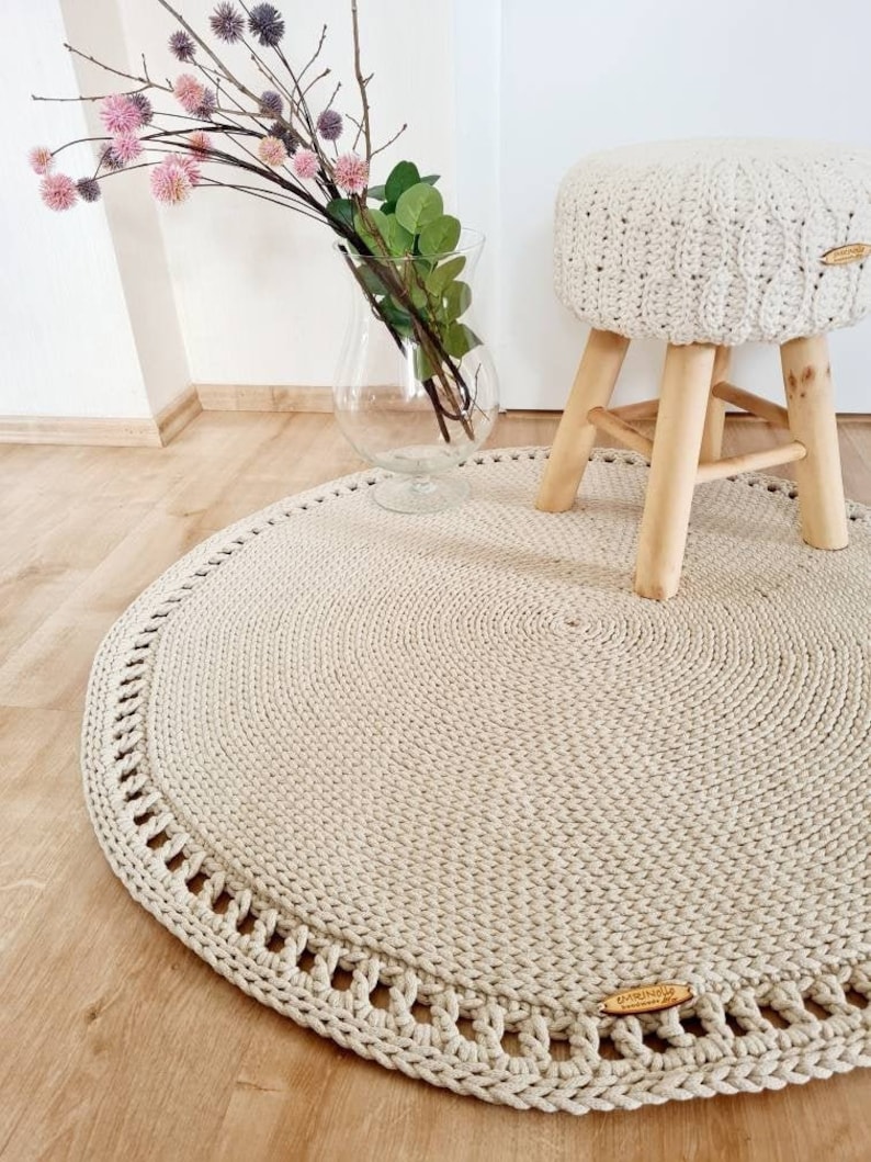 May include: A beige crocheted round rug with a decorative border. The rug is on a wooden floor and a small wooden stool with a beige crocheted seat is on top of the rug.