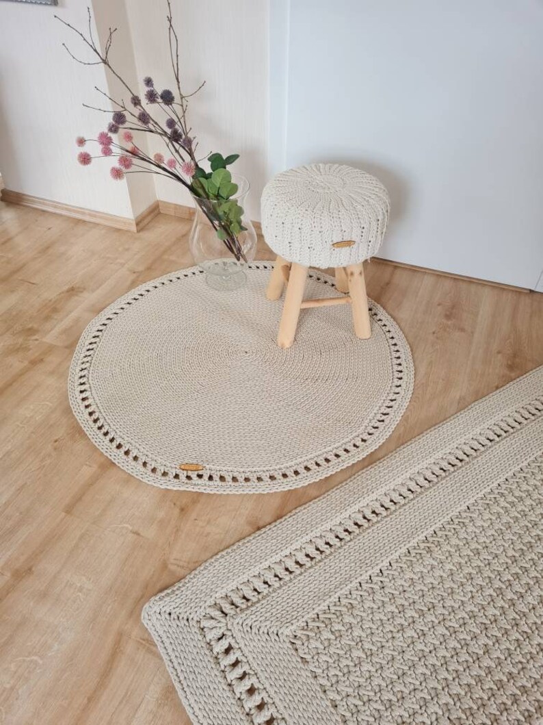May include: Two crocheted rugs in a beige color. One rug is round and the other is rectangular. A small wooden stool with a crocheted cushion is sitting on the round rug.