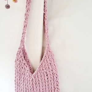 Puede incluir: Una bolsa de mano de crochet rosa con una correa trenzada. La bolsa est&aacute; colgada en una pared blanca.