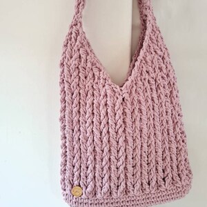 Puede incluir: Un bolso de mano de crochet rosa con un bot&oacute;n de madera. El bolso tiene un dise&ntilde;o trenzado y un fondo ancho y plano.