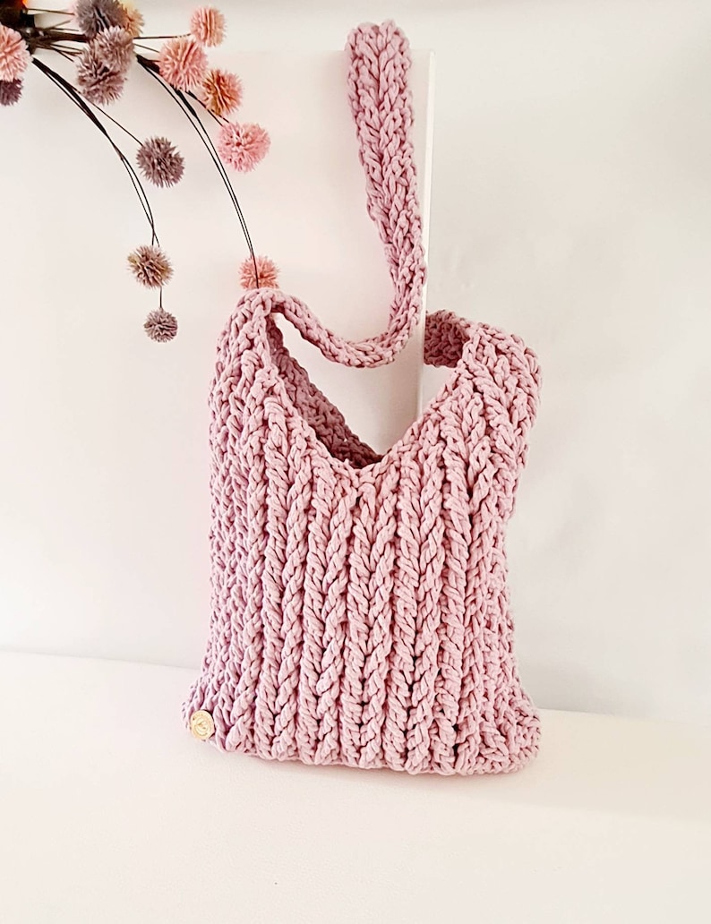 Puede incluir: Una bolsa de mano de crochet rosa con una sola correa. La bolsa est&aacute; hecha con un punto texturizado y tiene un peque&ntilde;o bot&oacute;n dorado en el lateral.