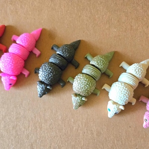 May include: A collection of colorful, segmented lizard-shaped toys. The toys are in various colors, including red, pink, gray, green, and white. Each lizard has a textured body and small legs, with a pointed head.