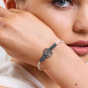 Puede incluir: Una pulsera plateada con un diseño de nudo celta. La pulsera presenta una banda plateada tejida y detalles en cuero gris oscuro. El nudo es de color plateado pulido. La pulsera se lleva en la muñeca.