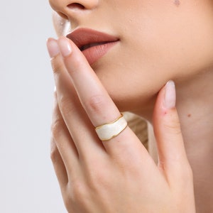 Könnte beinhalten: Ein weißer Ring mit einem goldenen Band. Der Ring hat eine wellige, organische Form.