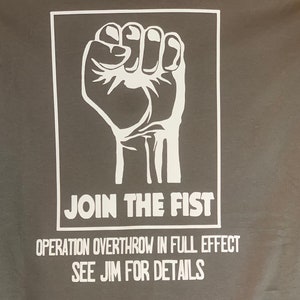 May include: Gray t-shirt featuring a white graphic of a clenched fist inside a white rectangle. Below the graphic, the text reads "JOIN THE FIST." Additional text below states "OPERATION OVERTHROW IN FULL EFFECT SEE JIM FOR DETAILS."