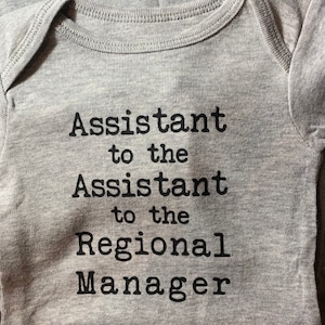 May include: A heather gray baby bodysuit featuring black text printed on the front that reads: Assistant to the Assistant to the Regional Manager. The garment has short sleeves and a ribbed crew neckline, with a portion of another garment visible.