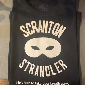 May include: Black t-shirt with the words "SCRANTON STRANGLER" in white, above a white mask graphic. Below the text is the phrase "He's here to take your breath away."