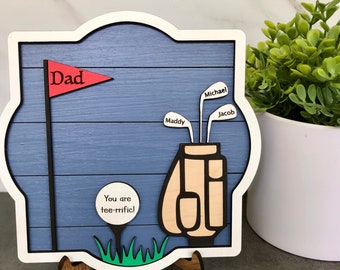 May include: A blue and white wooden sign with a golf bag, golf clubs, and a golf flag. The flag says "Dad" and the sign says "You are tee-rific!" The golf clubs have the names "Maddy", "Michael", and "Jacob" on them.