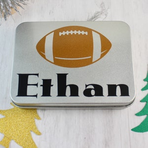 May include: A silver metal tin with a brown football graphic and the name "Ethan" printed in black.
