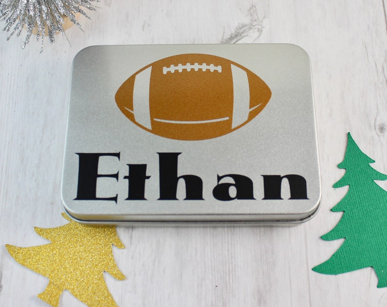 May include: A silver metal tin with a brown football graphic and the name "Ethan" printed in black.