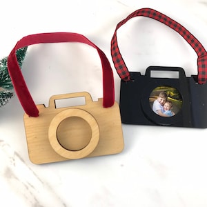 May include: Two wooden camera ornaments with red and black ribbon hangers. One camera is natural wood with a clear window for a photo. The other camera is black with a photo of two boys inside the window.
