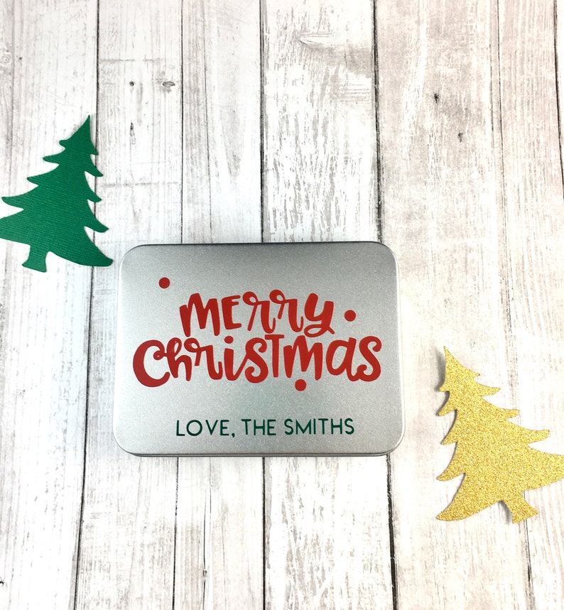 May include: A silver metal tin with a red "Merry Christmas" message and the text "Love, The Smiths" on a white wooden background with two paper Christmas trees.