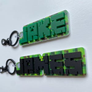 May include: Two keychains with the names "JAKE" and "JAMES" in pixelated block letters. The letters are green and black, respectively, and are set against a green and brown pixelated background. Each keychain has a black ring.