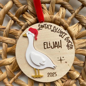May include: A wooden Christmas ornament featuring a white goose wearing a Santa hat. The ornament has the text "Santa's Lilliest Goose" and the name "Elijah" with the year "2025" below the goose. A red ribbon is attached.