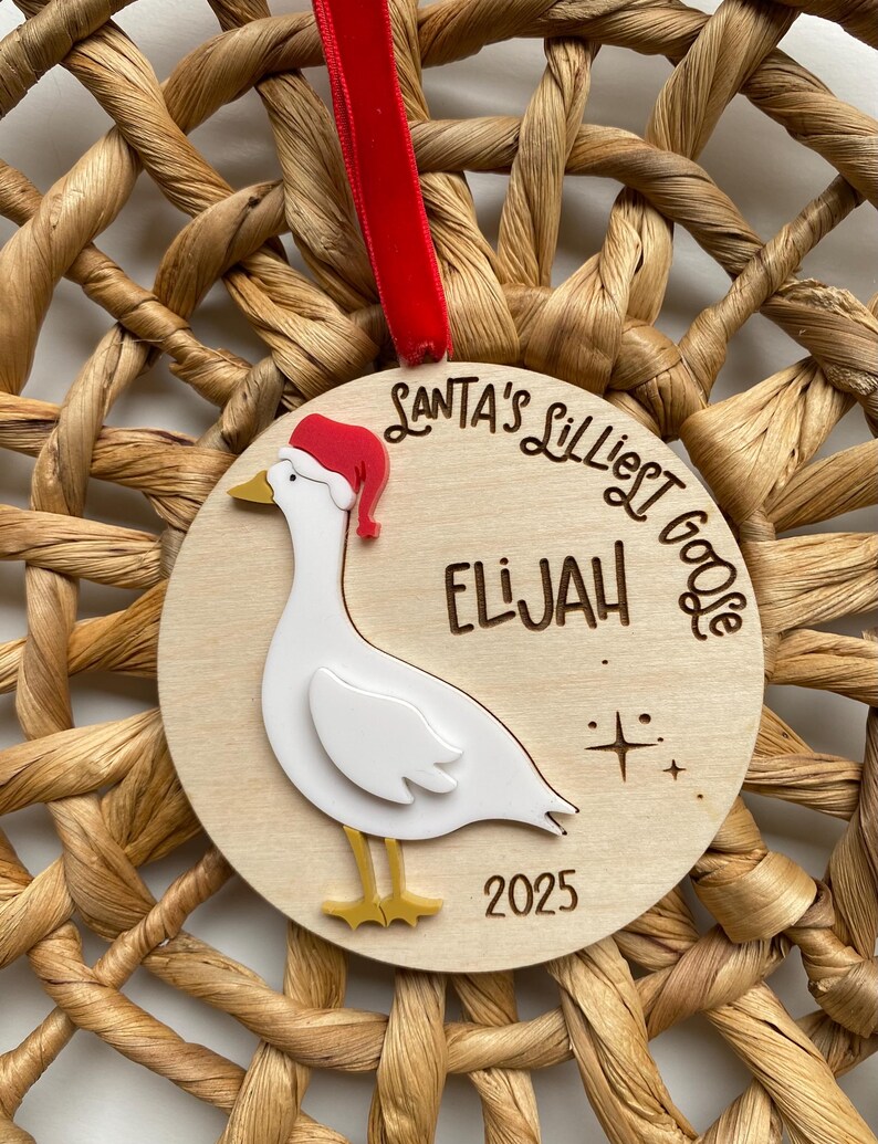 May include: A wooden Christmas ornament featuring a white goose wearing a Santa hat. The ornament has the text "Santa's Lilliest Goose" and the name "Elijah" with the year "2025" below the goose. A red ribbon is attached.