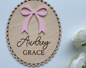 May include: A wooden oval sign with a pink bow and the name "Audrey Grace" in cursive. The sign has a scalloped edge and is made of light-colored wood. The bow is a soft pink color.