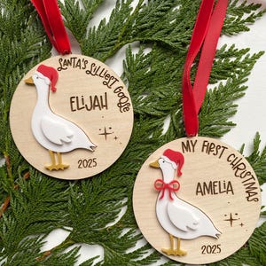 May include: Two wooden Christmas ornaments with red ribbon hangers. Each ornament features a white goose wearing a Santa hat. One says "Santa's Littlest Goose" and "Elijah 2025". The other says "My First Christmas" and "Amelia 2025".