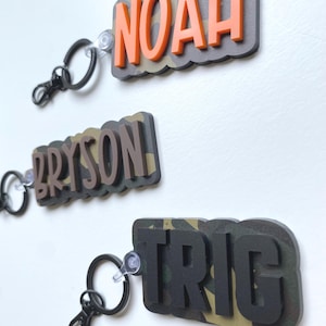 May include: Three personalized name keychains. Each keychain features a name in bold, raised letters. The names are "NOAH" in orange, "BRYSON" in brown, and "TRIG" in black. The background has a camouflage pattern.