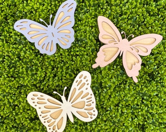 May include: Three wooden butterfly cutouts featuring intricate wing patterns with openwork details. One butterfly is light purple, one is soft pink, and one is a natural light wood color, each displaying a layered design with smooth edges.